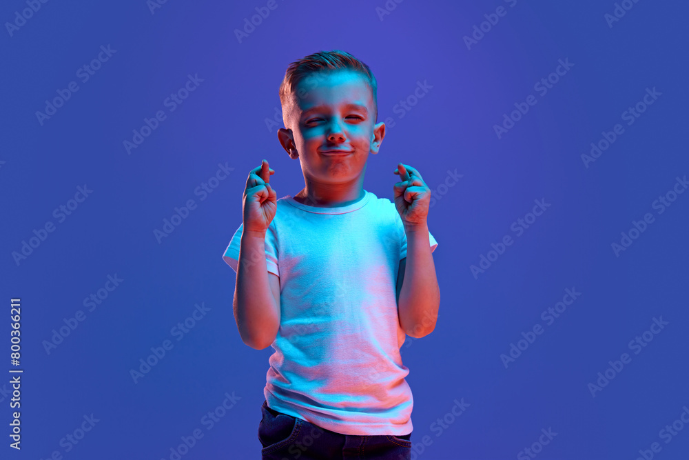 Good luck at exam. Cute child, boy crossed fingers and closed eyes to get lucky in mixed neon light against blue background. Concept of fashion and style, beauty, back to school, education, fortune. Good luck at exam. Cute child, boy crossed fingers and closed eyes to get lucky in mixed neon light against blue background. Concept of fashion and style, beauty, back to school, education, fortune.