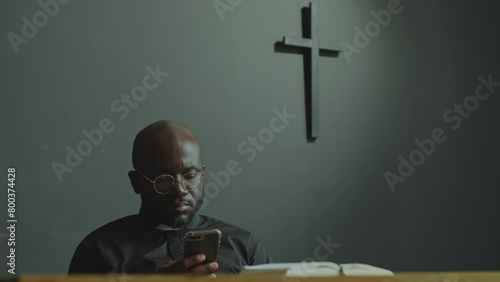 Black Catholic priest in cassock and glasses sitting in modern church and reading news on the Internet on mobile phone photo