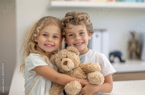 A young boy and girl smile while holding a teddy bear together in a doctor's office or hospital clinic