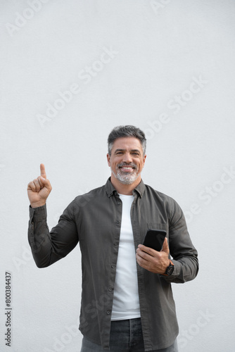 Mature business man using smartphone mobile phone app, pointing up at copy space for promo advertising. Freelance entrepreneur holding cellphone isolated on white background, looking camera, vertical
