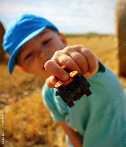 Happy 3-year old Caucasian boy showing a small tractor toy in stubble on a summer day. Concept for happy carefree childhood, enjoying childhood and summer, Children's Day. Concept for harvest end.