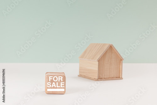 Simplistic wooden house model isolated on pale green background, with text For Sale on signboard.