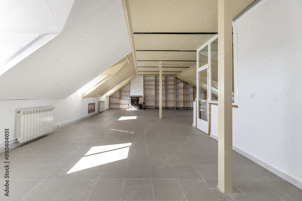 Large attic with a wall of shelves, windows with skylights, glass staircase, metal columns in the center, white aluminum radiators, fireplace and gray floor. Large attic with a wall of shelves, windows with skylights, glass staircase, metal columns in the center, white aluminum radiators, fireplace and gray floor.