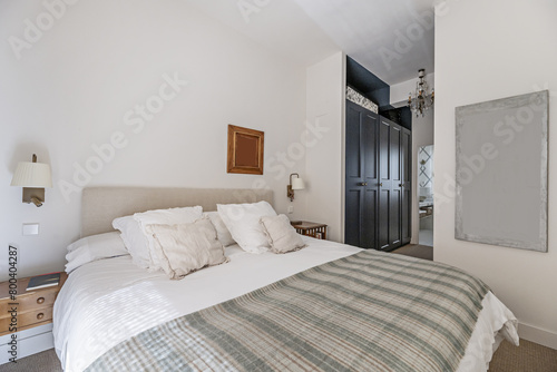 Double bedroom with fabric upholstered headboard and many cushions and pillows on the mattress, a hallway with a walk-in wardrobe and access to a bathroom