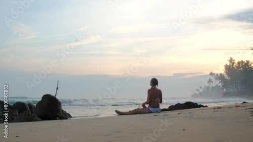 Woman silhouette practicing sitting straddle on sandy beach near ocean in mist. Lady athlete enjoys yoga session at summer sunset photo