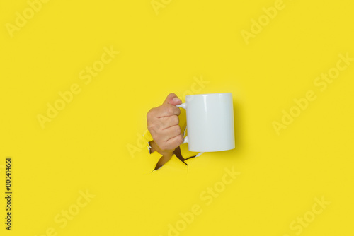 Female hand coming out of a hole in a torn yellow paper background holding white cup. Mockup.