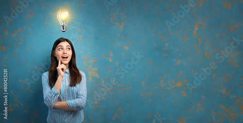 Illuminated light bulb above the head of a young woman, the concept of having an idea of a surprised girl with eureka on an old blue wall 