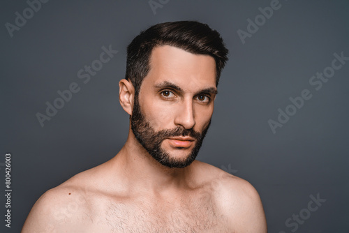 Attractive young shirtless man with beard looking at the camera isolated over grey background. Sexy naked male model with hairstyle posing for barbershop advertisement