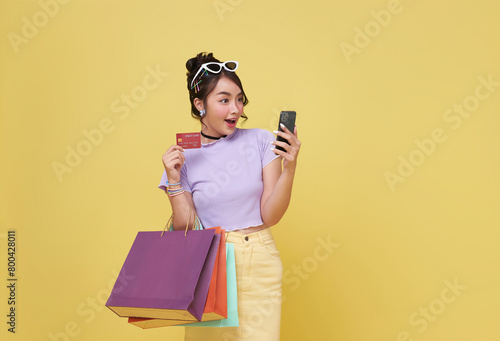 Excited Asian woman carrying shopping bags with credit card and looking mobile phone in hands isolated on yellow background.