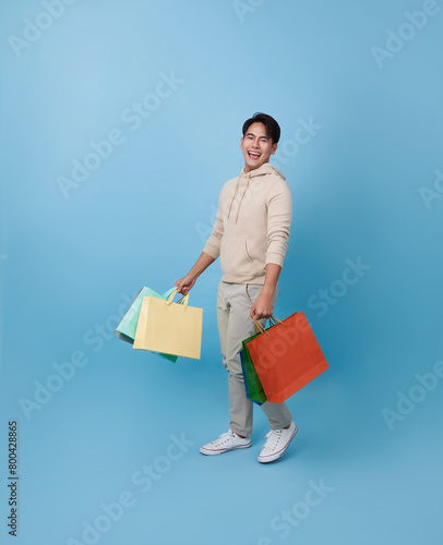Full length young asian shopper man walking and holding shopping bag feeling happy excited for discount promotion isolated on blue background.