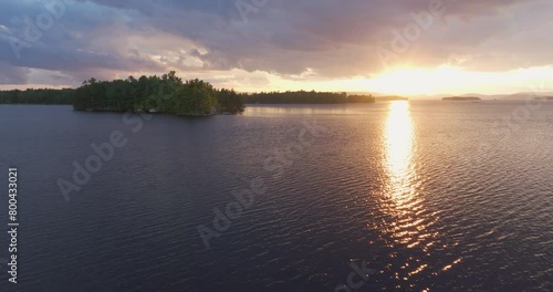 From Above drone footage of rippled Ambajejus Lake with trees and sunlight reflections under overcast dramatic sky during sunset in Maine