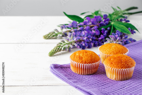 Soft fresh muffins on textile napkin. Delicious pastries and purple lupine flowers on white wooden background. Preparing dessert in confectionery. Copy space. © Galina Atroshchenko