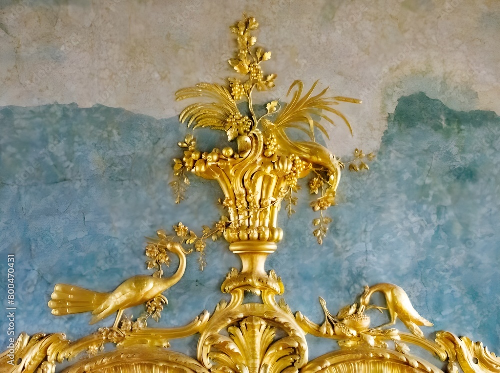 A gold decoration featuring a basket of flowers and fruit, a crane, and a peacock, is mounted on a teal wall.