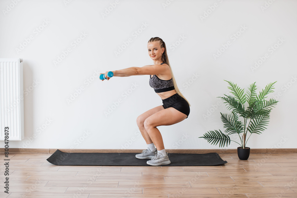 A focused young woman in athletic wear engages in a squat exercise with dumbbells in a bright, minimalist room with a plant. A focused young woman in athletic wear engages in a squat exercise with dumbbells in a bright, minimalist room with a plant.