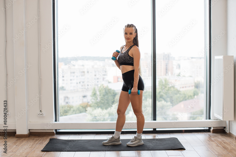 A fit young woman holds blue dumbbells, ready to workout in a bright home environment. She stands on a yoga mat by a large window overlooking the city.