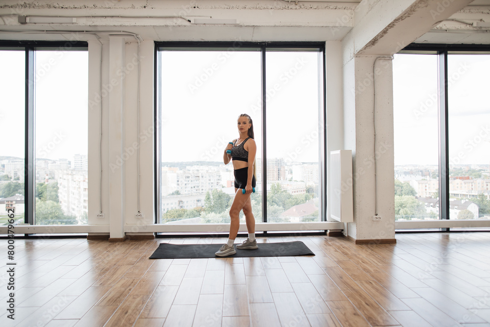 Fit young woman takes break during an intense workout in spacious modern gym with large windows overlooking city. Sports woman stands on yoga mat, demonstrating commitment to fitness and health. Fit young woman takes break during an intense workout in spacious modern gym with large windows overlooking city. Sports woman stands on yoga mat, demonstrating commitment to fitness and health.