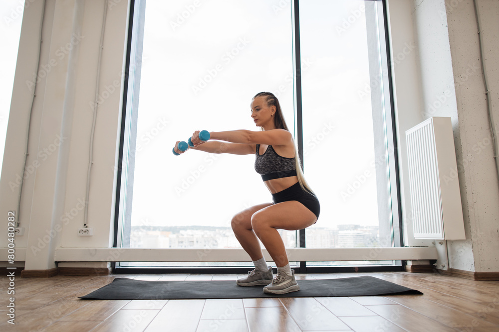 Fototapeta premium Focused young female athlete performs squats while holding dumbbells in a well-lit fitness center with large windows overlooking the city. Dedication and health consciousness in an urban setting.