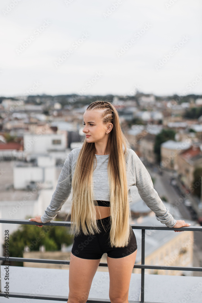 A young woman stands on a rooftop dressed in stylish athletic wear, overlooking a panoramic cityscape. Her relaxed posture and thoughtful expression reflect a moment of peace amidst urban life. A young woman stands on a rooftop dressed in stylish athletic wear, overlooking a panoramic cityscape. Her relaxed posture and thoughtful expression reflect a moment of peace amidst urban life.
