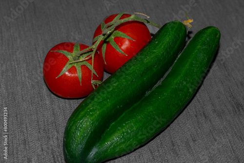 two fresh red tomatoes with stem still attached and two fused cucumbers on a grey background