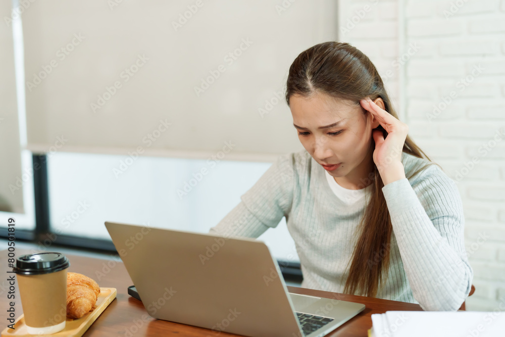 Beautiful young Asian woman Coffee shop owner sitting in front of a work desk in front of a laptop computer. Check sales work from notebook Hands gently massage the head showing a stressed expression Beautiful young Asian woman Coffee shop owner sitting in front of a work desk in front of a laptop computer. Check sales work from notebook Hands gently massage the head showing a stressed expression