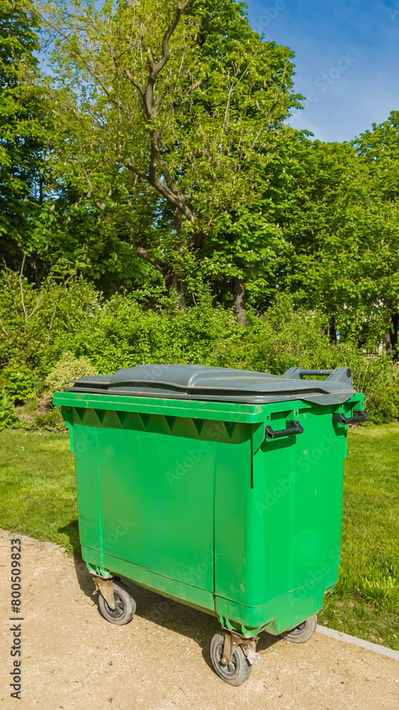 Green waste container on wheels in a sunny park, symbolizing urban recycling and environmental conservation efforts Related to: Earth Day, World Environment Day Green waste container on wheels in a sunny park, symbolizing urban recycling and environmental conservation efforts Related to: Earth Day, World Environment Day