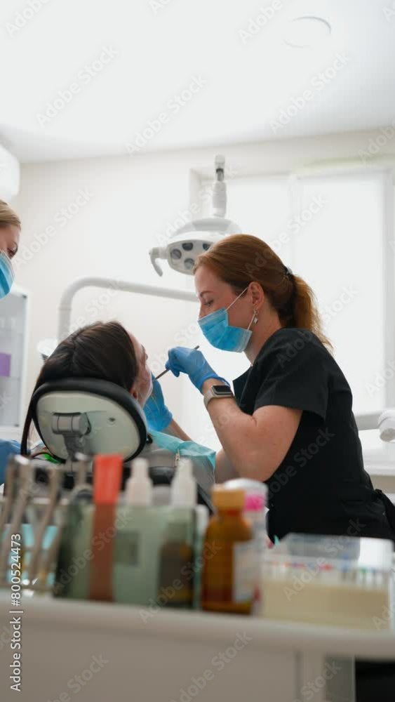 Two Young Adult Female Dentists in Uniform and Medical Masks Examine a Patient in a Modern Dental Clinic. Healthcare and Medicine Concept. Teeth, Caries and Dentistry. Slow Motion, Vertical Video. Two Young Adult Female Dentists in Uniform and Medical Masks Examine a Patient in a Modern Dental Clinic. Healthcare and Medicine Concept. Teeth, Caries and Dentistry. Slow Motion, Vertical Video.