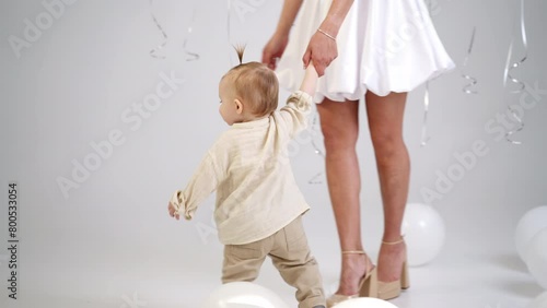 Unrecognized woman in short white dress holding her baby by the hand. Cute smiling child walks around his momma. Studio footage with white balloons around. photo