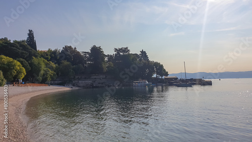 A small pier along the shore of Medveja in Croatia. The Mediterranean Sea is calm and clear. There is a lush forest with a small town at the shore. Clear, blue sky. Summer remedy. Stony beach. photo