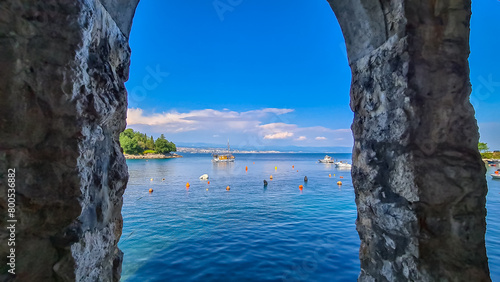 A view through a stony arch on the cities located along the coastal line of Istria, Croatia. There are plenty of buoys in the water and a few boats parked in there. The Mediterranean Sea is calm.