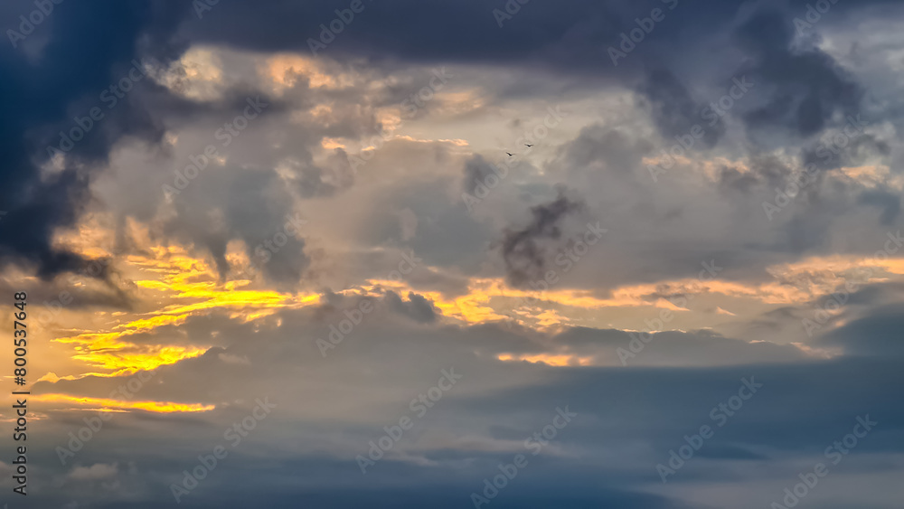 The setting sun hiding behind thick clouds. The clouds are colored yellow and orange. Many different layers of the clouds. Calmness and quietness.