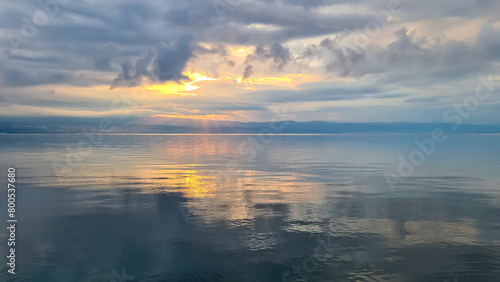 The sun setting behind mountains in Croatia. The Mediterranean Sea is calm and clear, reflecting the sunbeams. The sky is orange and covered with clouds. Many cities along the coastal line. Calmness