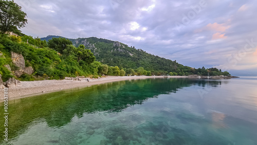 A stony beach along the shore of Medveja in Croatia. The Mediterranean Sea is calm and clear. There is a lush forest with a small town at the shore. The sky is painted yellow. Sunset time. Calmness photo