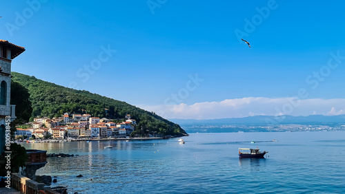 A panoramic view of the shore along Moscenicka Draga, Croatia. There is a small town located on the shore of the Mediterranean Sea. Few boats crossing the calm sea. Green hills in the back. Sunny day. photo
