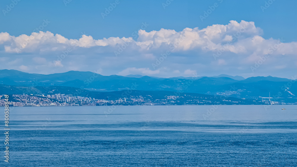 A panoramic view of the Croatian shore. There are some towns located on the shore of the Mediterranean Sea. Calm surface of the sea. Stony beach. Green hills in the back. Sunny day with puffy clouds