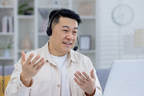 A cheerful Asian man engages in a lively conversation using a headset in a bright, neatly organized home office setting, demonstrating remote work communication.