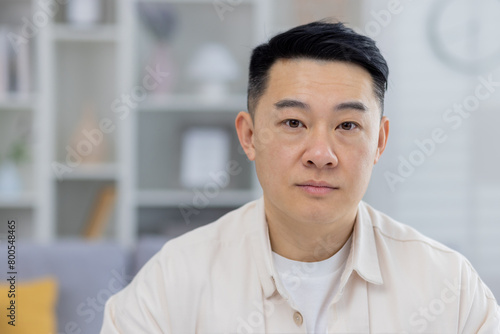A clear, high-quality photo of a middle-aged Asian man looking directly at the camera with a calm expression, set against a softly blurred home background.