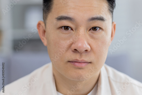 Fotografie A detailed close-up portrays a middle-aged Asian man with a contemplative, serious expression, captured indoors with a soft focus background