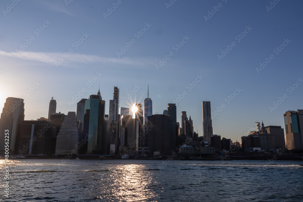 Fototapeta premium Manhattan cityscape at dusk with iconic skyscrapers reflecting on the Hudson River. Great density of skyscrapers, some of them are reflecting the sunbeams of setting sun. Vibrant city.