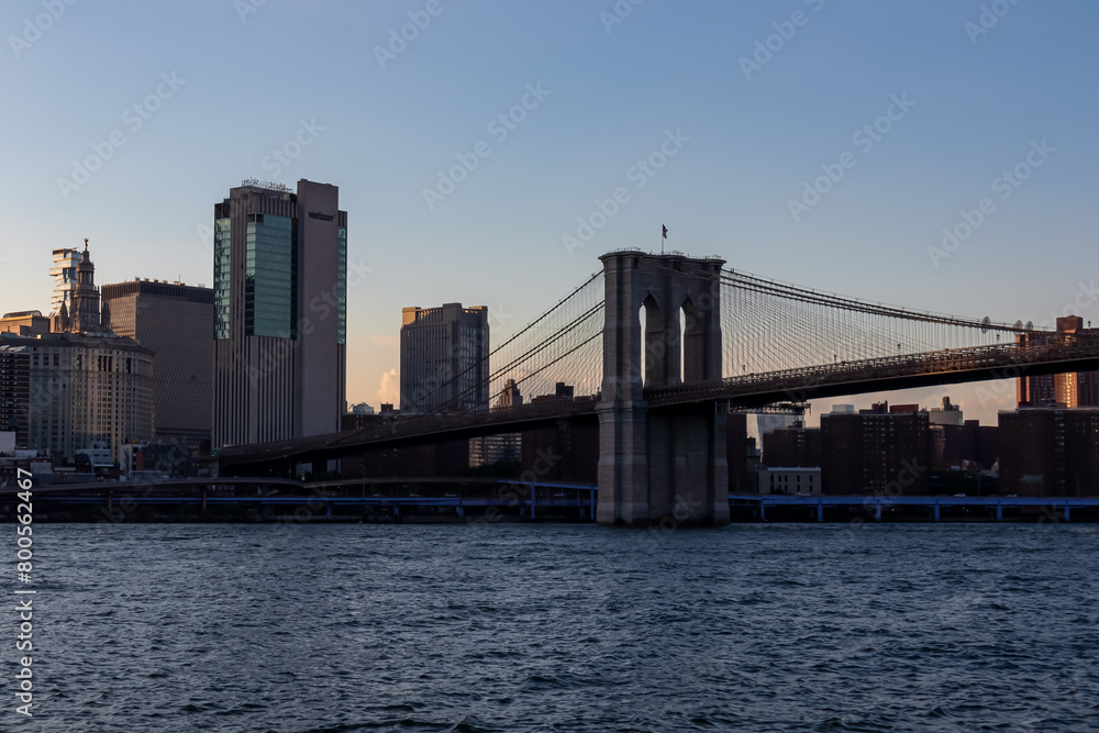 Naklejka premium Iconic Brooklyn Bridge connecting New York City's urban landscape seen from below. There are a lot of lush trees in the frame. Clear blue sky above. Sightseeing in the city. Summer vibes