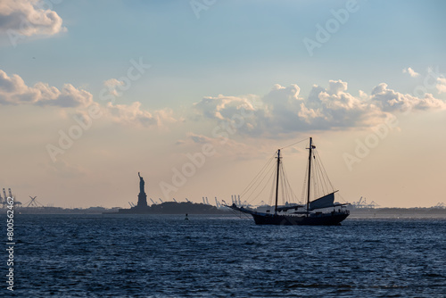 A sailing boat crossing the bay of New York with the view on Statue of Liberty in the back. There are a lot of factories over the horizon line. Few puffy clouds on the sky. Gentle colors of sunset. photo