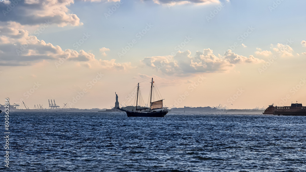 A sailing boat crossing the bay of New York with the view on Statue of Liberty in the back. There are a lot of factories over the horizon line. Few puffy clouds on the sky. Gentle colors of sunset.