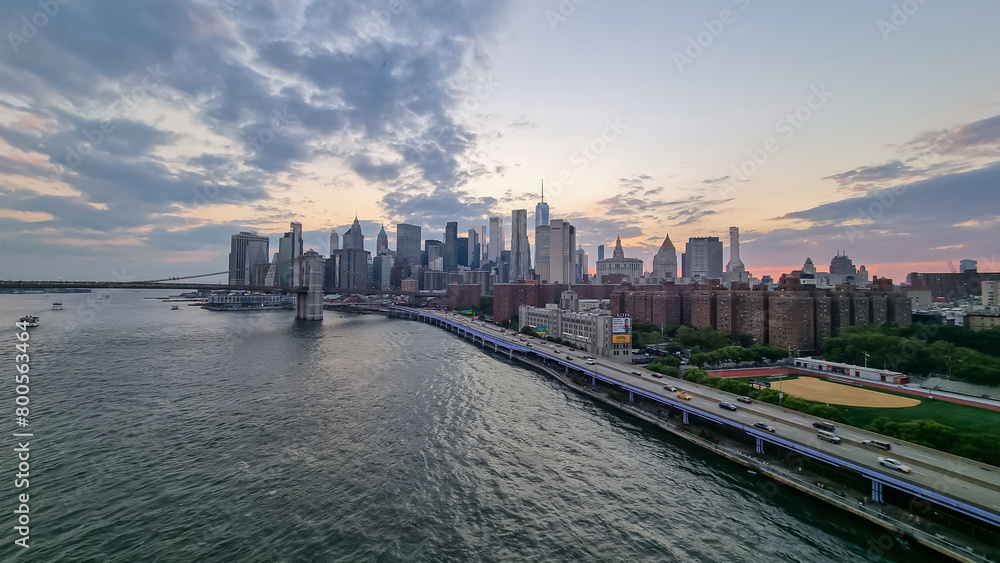 Fototapeta premium A view at the panorama of New York City's skyscrapers. Brooklyn Bridge in front is lit up. Overcast. Missing freedom. The sun is slowly setting behind the city. Busy and dazzling city.