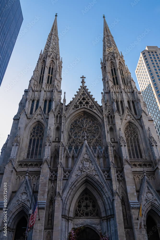 St. Patrick Cathedral in New York City seen from the street across. The Cathedral is built between high skyscrapers. Clear and blue sky above it. American flag attached to the cathedral. St. Patrick Cathedral in New York City seen from the street across. The Cathedral is built between high skyscrapers. Clear and blue sky above it. American flag attached to the cathedral.