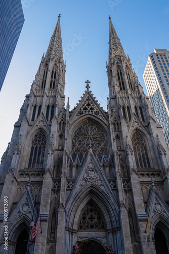 St. Patrick Cathedral in New York City seen from the street across. The Cathedral is built between high skyscrapers. Clear and blue sky above it. American flag attached to the cathedral.
