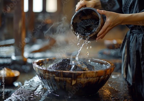 Close up of a woman's hands pouring water from a wooden bucket into hot stones in a Japanese onsen steam room. Wood walls and floor with steam rising. A large bowl filled with black rocks. 