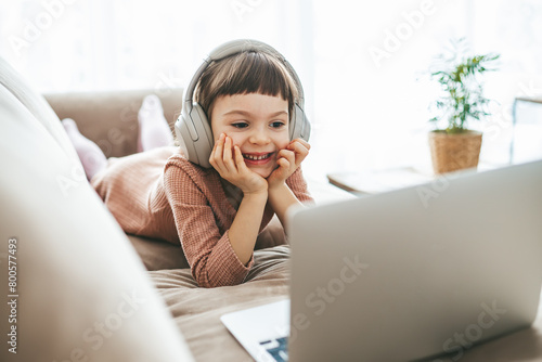 With headphones on, a sweet little girl, relaxing on a sofa, engrossed in the captivating content playing on her laptop