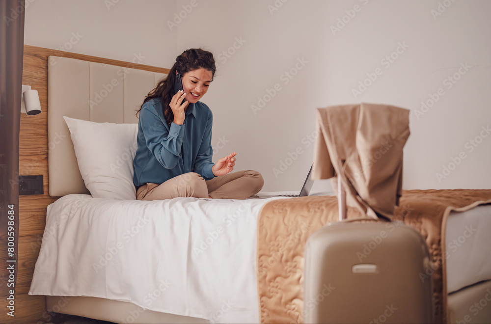 Businesswoman sitting on bed, using laptop talking over phone. Young modern woman executive on business trip working in hotel room. Businesswoman sitting on bed, using laptop talking over phone. Young modern woman executive on business trip working in hotel room.