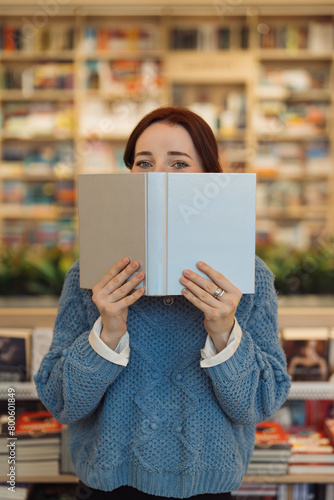 A young red-haired woman covers her face with a book in a cozy library setting, surrounded by shelves filled with books.