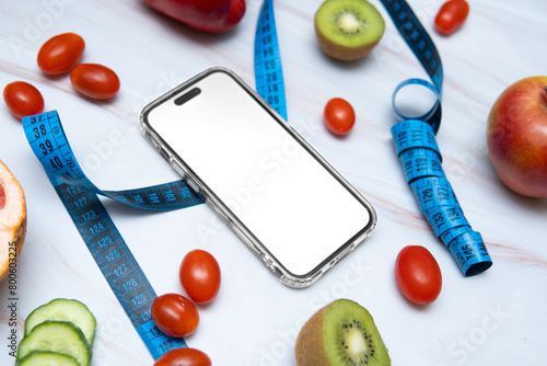 A smartphone mockup placed next to a tailor's measuring tape and an assortment of healthy fruits and vegetables, including cucumbers, peppers, tomatoes, kiwi, and apples.