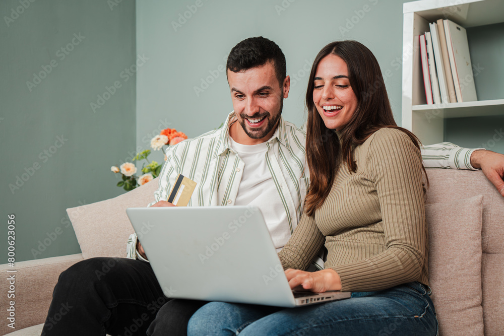 Happy young adult caucasian couple browsing on internet using a laptop to search a sale at home. Excited husband and wife smiling and looking for new apartment sitting on a sofa at living room